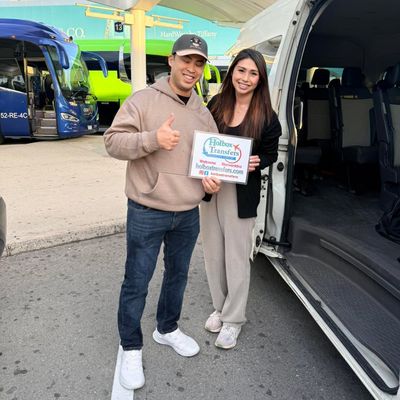 Pareja sonriente de pie junto a la puerta abierta de la van sostiene el banner de Holbox Transfers, preparándose para viajar a Holbox.