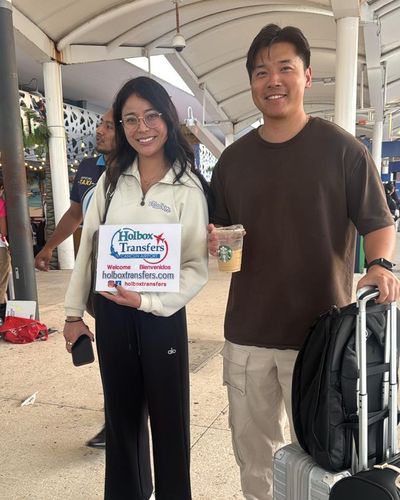 Pareja sonriente de pie en la terminal con café en mano, sosteniendo el banner de Holbox Transfers antes de su traslado a Holbox.