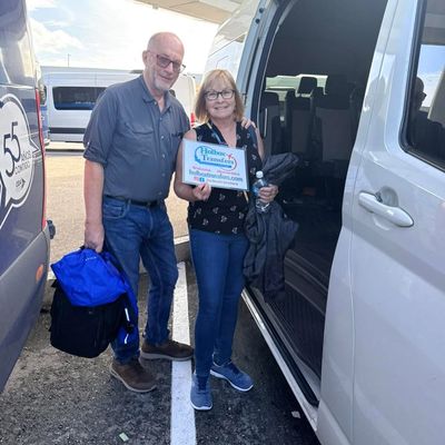 Pareja sonriente de pie en el área de estacionamiento de la terminal, sosteniendo el banner de Holbox Transfers antes de su traslado a Holbox.