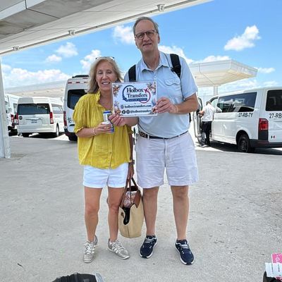 Pareja sonriente de pie en la terminal del aeropuerto, sosteniendo en alto el banner de Holbox Transfers antes de salir hacia Holbox.