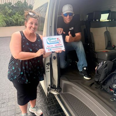 Pareja sonriente de pie junto a la van sostiene el banner de Holbox Transfers, saludando al conductor para el traslado Aeropuerto de Cancún - Holbox.