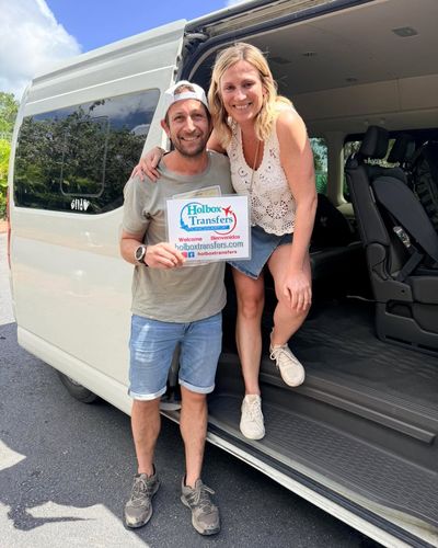 Pareja sonriente de pie junto a la puerta abierta de la van sostiene el banner de Holbox Transfers, lista para el traslado Aeropuerto de Cancún - Holbox.