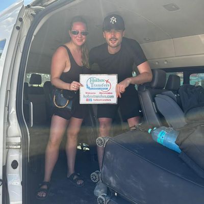 Pareja sonriente de pie dentro de la van sostiene el banner de Holbox Transfers, lista para su traslado entre el Aeropuerto de Cancún y Holbox.