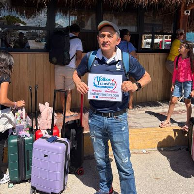Un viajero solo sostiene el banner de Holbox Transfers afuera de una terminal con techo de palapa, recién llegado a la isla de Holbox.