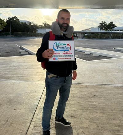 Un viajero solo sostiene en alto el banner de Holbox Transfers en la soleada terminal del aeropuerto, esperando salir hacia Holbox.