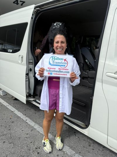 Viajero solo sonríe sosteniendo el banner de Holbox Transfers junto a la puerta abierta de la van antes de salir hacia Holbox.